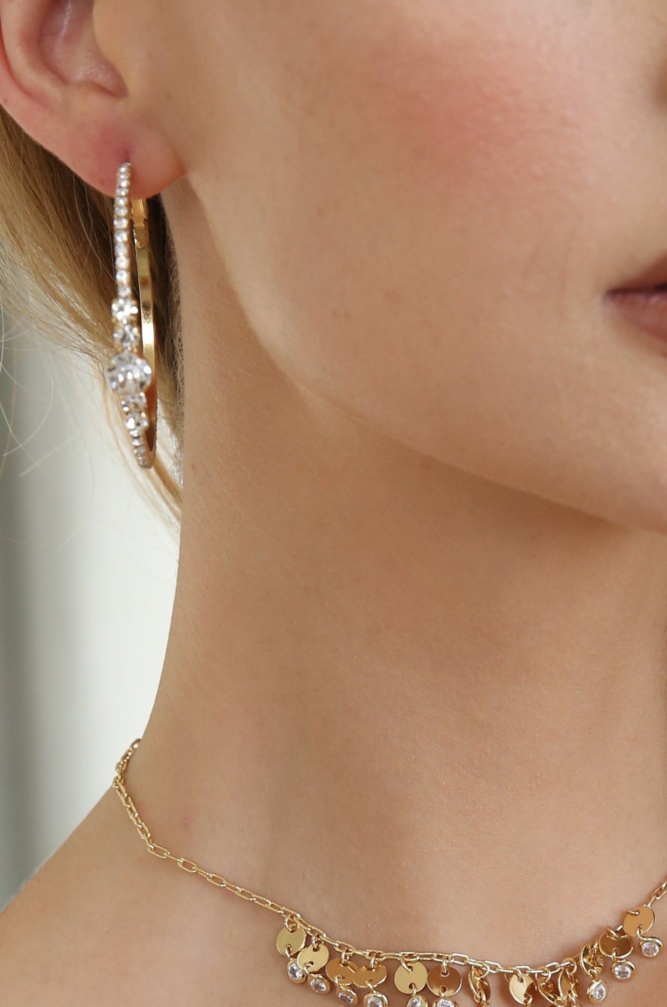 A close-up of a womanâ€™s neck features a gold necklace with a pendant and a large, ornate gold hoop earring adorned with sparkling stones, set against a light background.