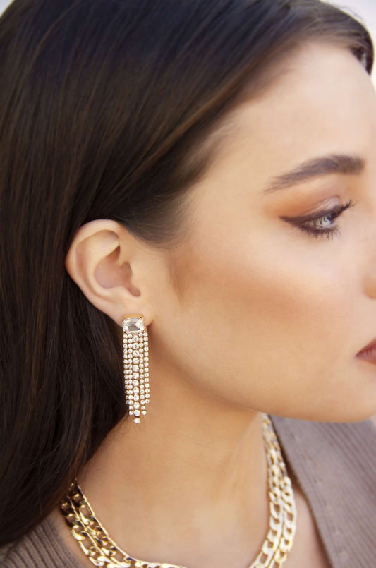 A long, sparkling earring hangs from a woman's ear, shimmering with multiple chains of rhinestones. She has long, dark hair and is wearing a stylish outfit, with a gold necklace visible.