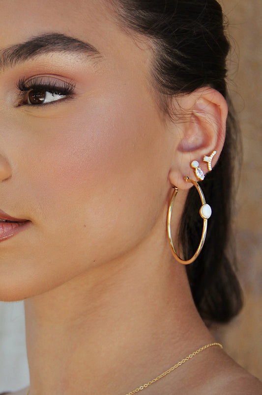 A young woman displays intricate earrings, including a large gold hoop and studded designs, set against a softly blurred, neutral background that emphasizes her facial features and delicate jewelry.