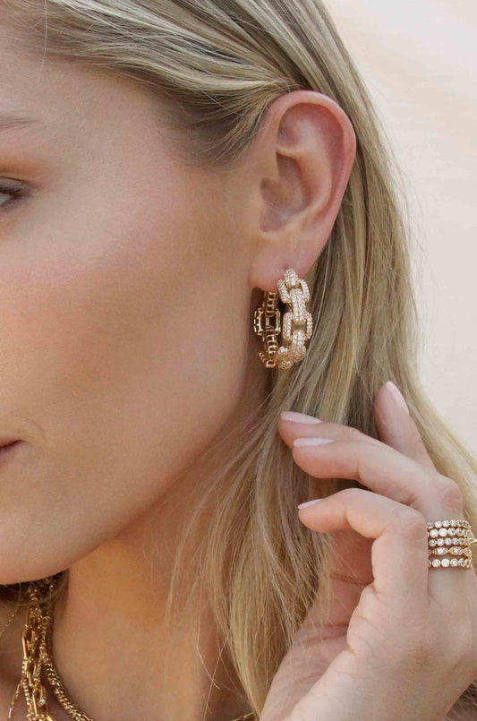 A close-up of a person wearing elaborate gold hoop earrings. They are gently touching one earring with their hand, set against a softly blurred background.