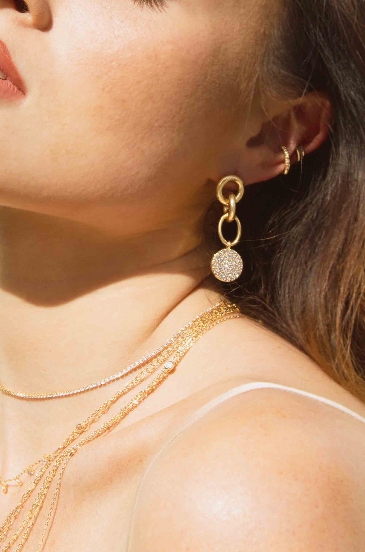 A person's neck and shoulder are adorned with layered gold necklaces. A large gold earring with a sparkling circular detail hangs from one ear against a softly lit background.