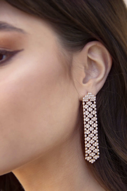 A sparkling, detailed chandelier earring hangs from a woman's ear. Her face is turned slightly, showcasing smooth skin and a soft makeup look, with hair cascading nearby.