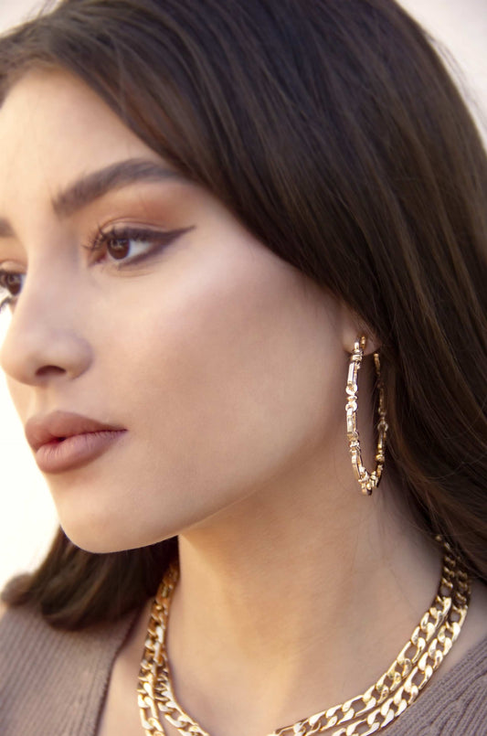 A young woman with long hair displays large gold hoop earrings and layered gold necklaces. She poses gracefully, showcasing her makeup, particularly the bold eyeliner, against a soft, neutral background.