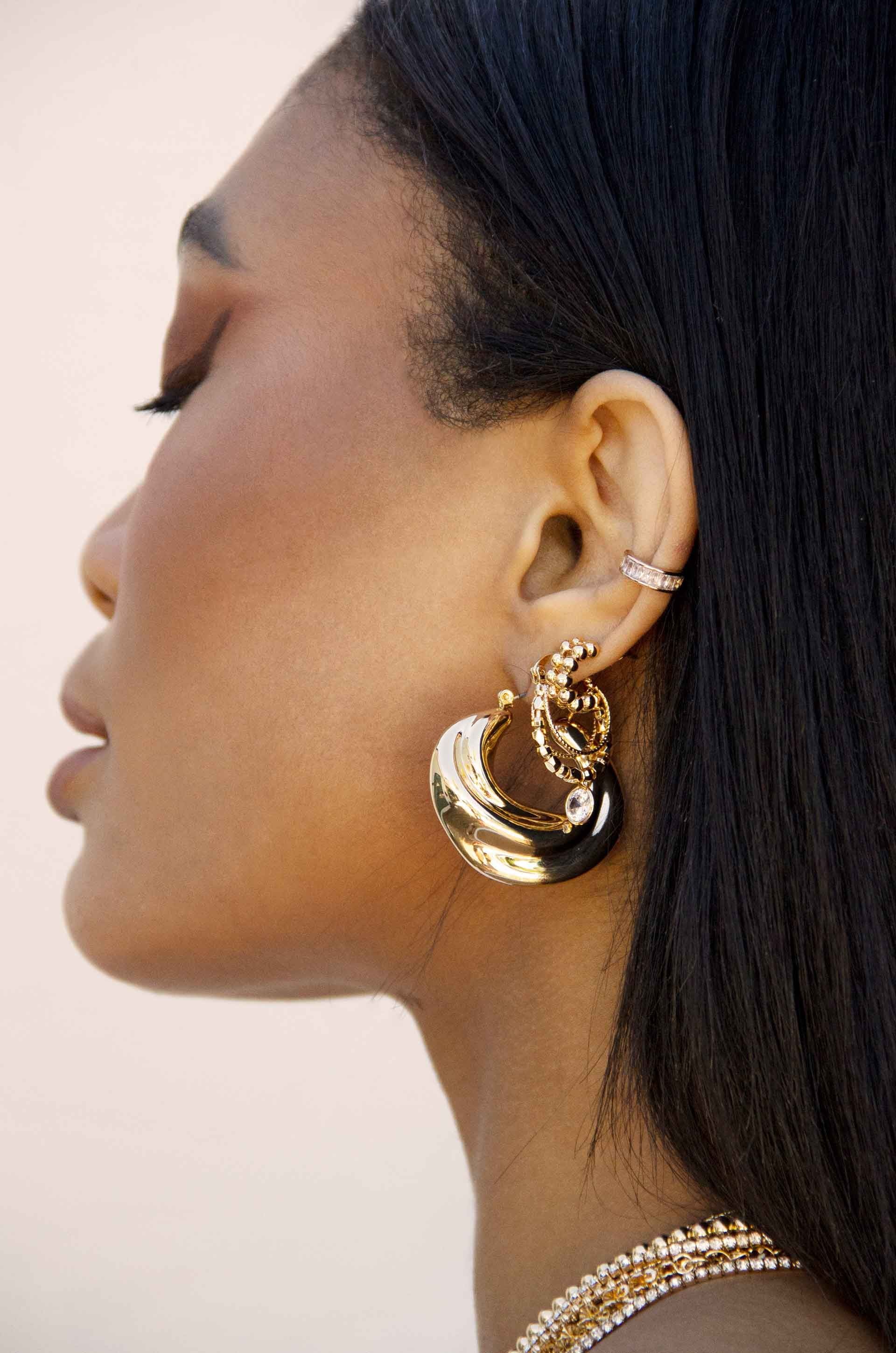 A woman with sleek black hair wears eye-catching gold and black earrings. She turns her head slightly, revealing her earlobe adorned with a small diamond-studded hoop. The background is softly blurred.