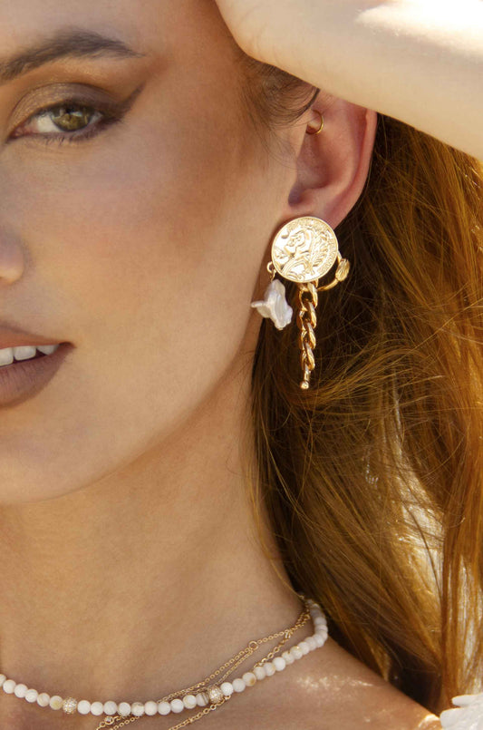 An elegant woman displays a unique gold earring with a round, intricate design and cascading elements, alongside layered necklaces, in a bright outdoor setting that highlights her features.