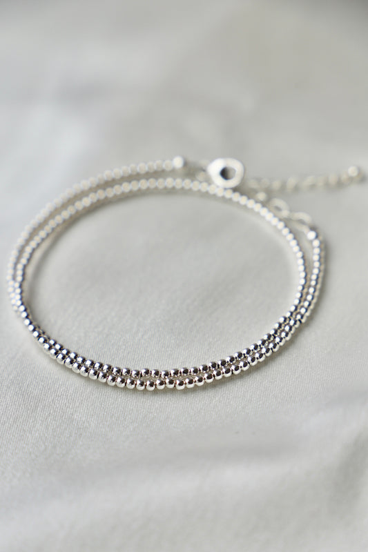 A silver bracelet with two intertwined strands of small beads rests on a softly illuminated, textured fabric surface, emphasizing its shine and delicate design.