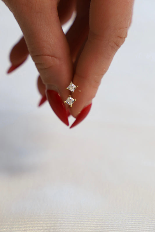 A hand with long red nails holds a pair of gold earrings featuring two square, clear gemstones, against a blurred, light-colored background.