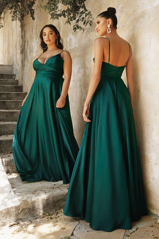 Two women wear elegant, green evening gowns, one facing forward and the other in profile. They stand on stone steps against a textured wall, with leaves above adding a natural touch.