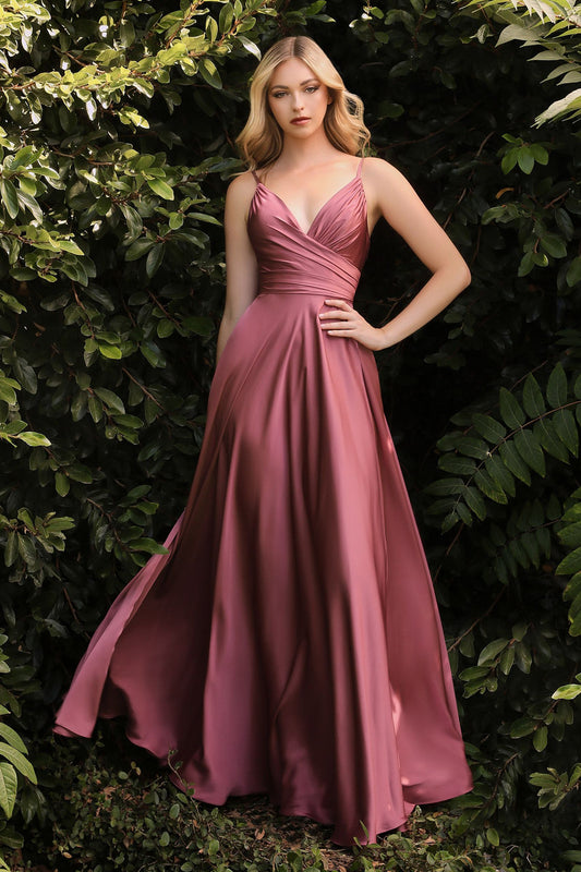 A woman stands confidently in a flowing, pink satin dress, with her hand on her hip, surrounded by lush green foliage, creating an elegant and serene atmosphere.