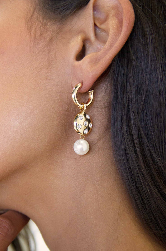 A gold earring with a round gem and a pearl hangs from a woman's ear, showcasing her smooth skin and dark hair. The setting is intimate and personal.