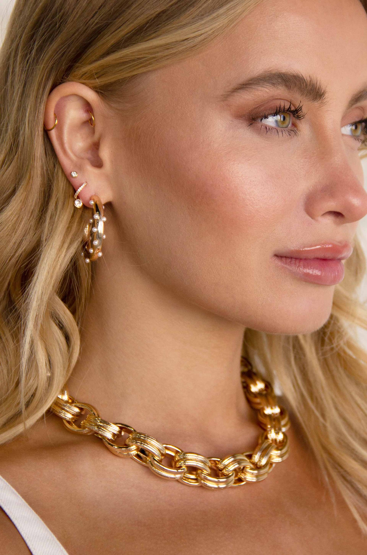 A woman with long, wavy blonde hair wears a large gold chain necklace and hoop earrings, showcasing a close-up profile against a soft, neutral background, highlighting her elegant features.