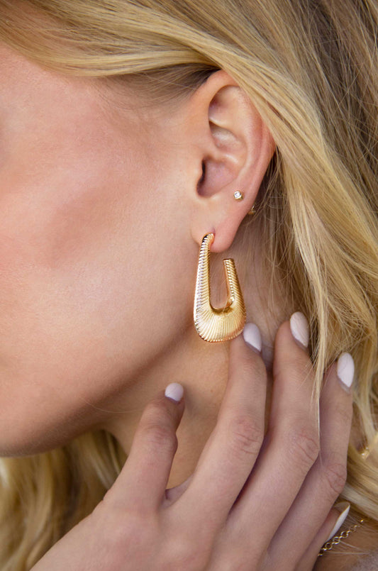 A large, gold-colored hoop earring hangs from a woman's ear. She touches the earring gently with her fingers, set against her blonde hair and skin, suggesting an intimate moment.