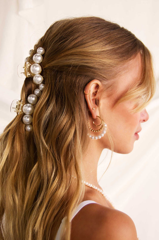 Long, flowing blonde hair is styled with pearl hair clips, enhancing elegance. The person wears multiple pearl and gold earrings, set against a soft, light background.