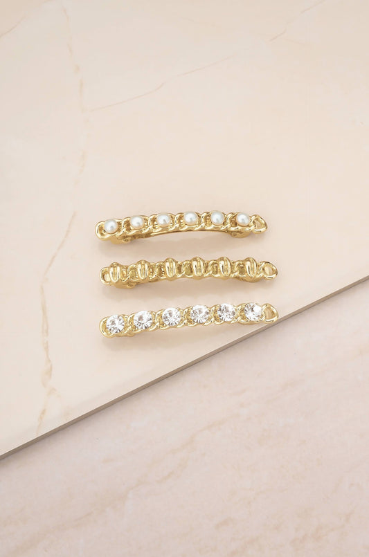 Three gold hair clips are arranged in a row on a light-colored, smooth surface. The clips feature pearls and clear gemstones, showcasing a decorative design suitable for hair styling.