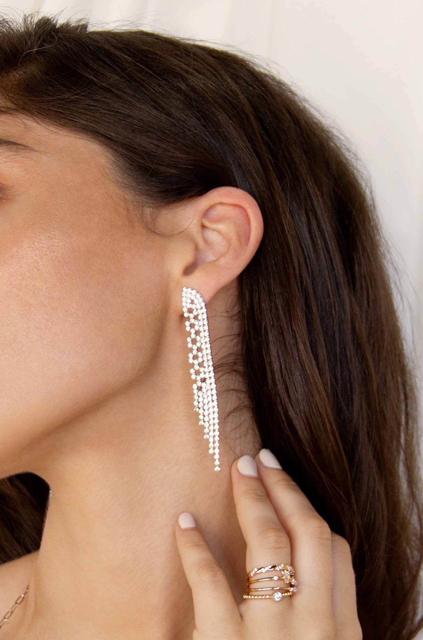 A woman wearing an elegant, long, beaded earring touches her neck with one hand, showcasing multiple rings, against a soft, light background. Her hair falls partially over her shoulder.