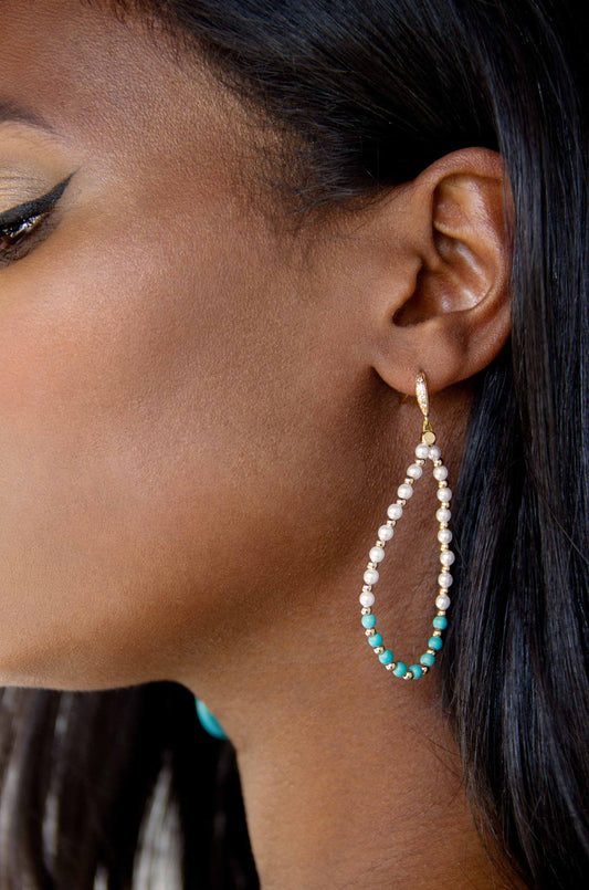 A teardrop earring made of alternating pearls and turquoise beads hangs decoratively from a woman’s ear, highlighting her smooth skin and long black hair against a neutral background.