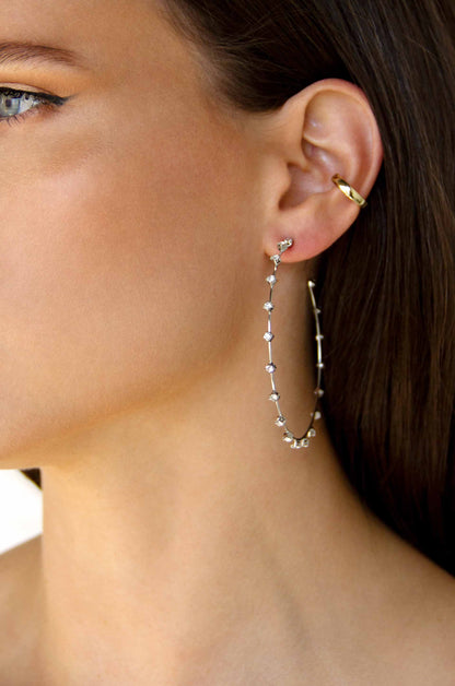 A close-up of a woman's ear features a long, delicate silver earring adorned with small gemstones, gently dangling beside a simple gold hoop earring. The skin tone appears soft and natural.