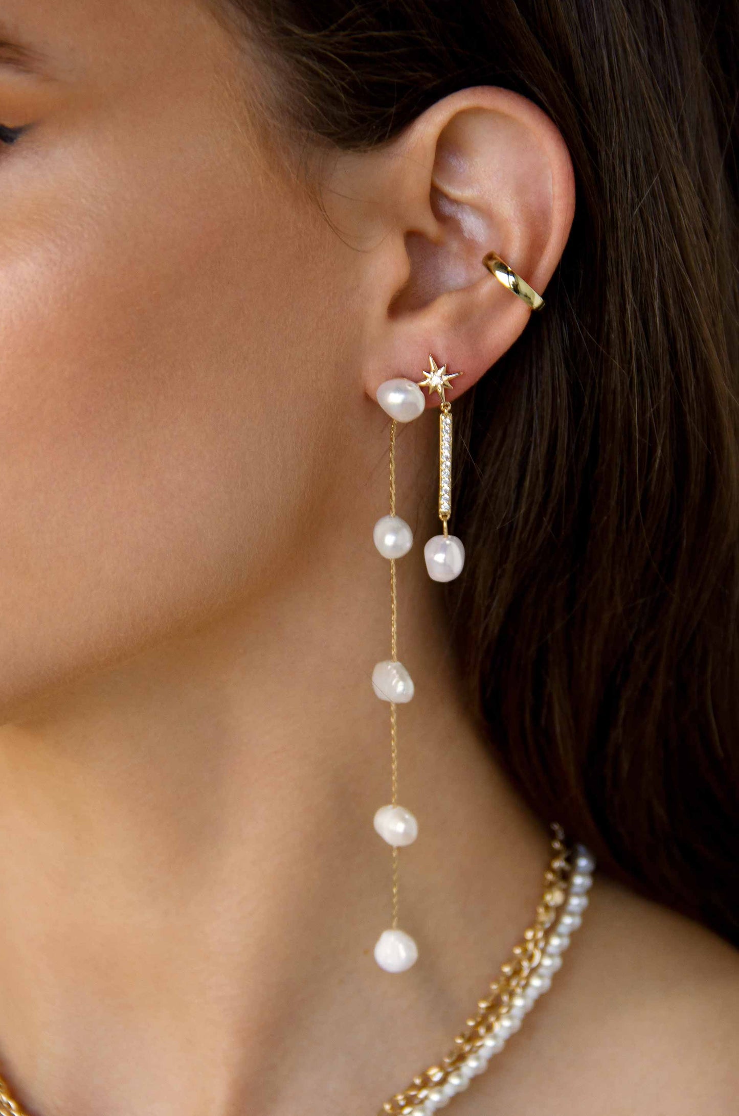 A close-up of a woman's ear showcases an intricate earring featuring a star-shaped top and a delicate chain adorned with white pearls, set against a softly illuminated, neutral background.