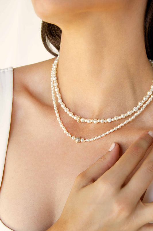 A person wears layered pearl necklaces. One hand gently touches the jewelry, highlighting the elegance of the pearls against smooth skin, set against a softly lit, neutral background.