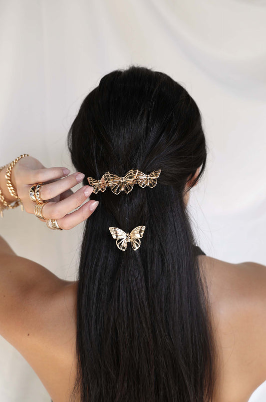 A hand holds a hair clip featuring multiple golden butterfly designs, positioned in long black hair. The background is soft and light, creating a gentle, elegant atmosphere.