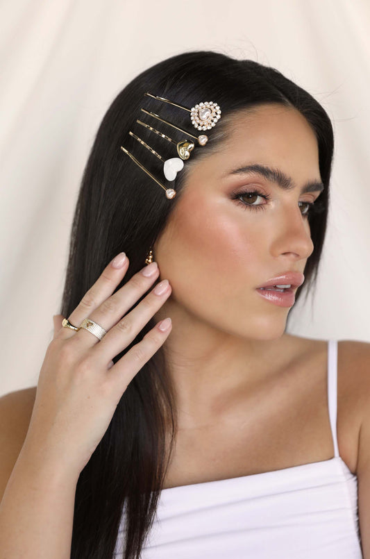 A woman with long, straight hair adorned with decorative hairpins touches her face gently. She wears a white sleeveless top and has a soft, neutral background that highlights her features.