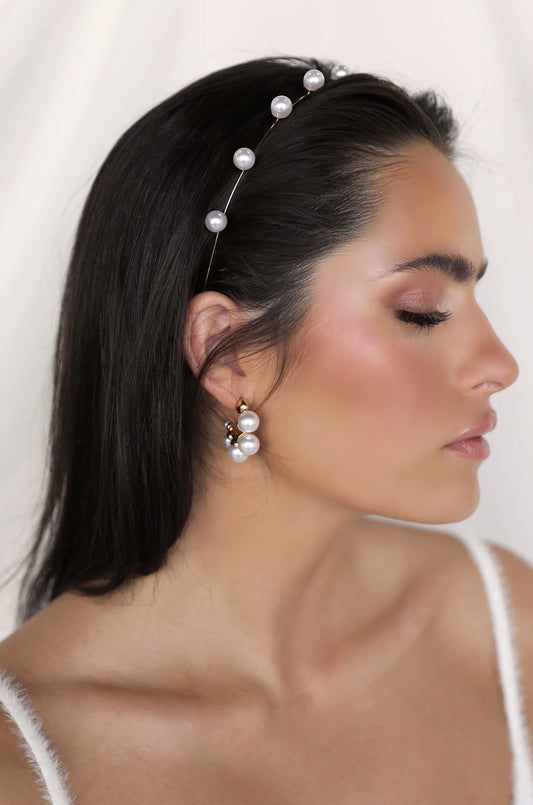 A young woman with long dark hair wears a pearl headband and pearl earrings. She gazes to the side against a soft, light background, emphasizing her elegant appearance.