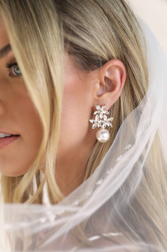 A close-up of a womanâ€™s ear adorned with a luxurious earring featuring sparkling gemstone accents and a large pearl, complemented by her flowing blonde hair and delicate veil.