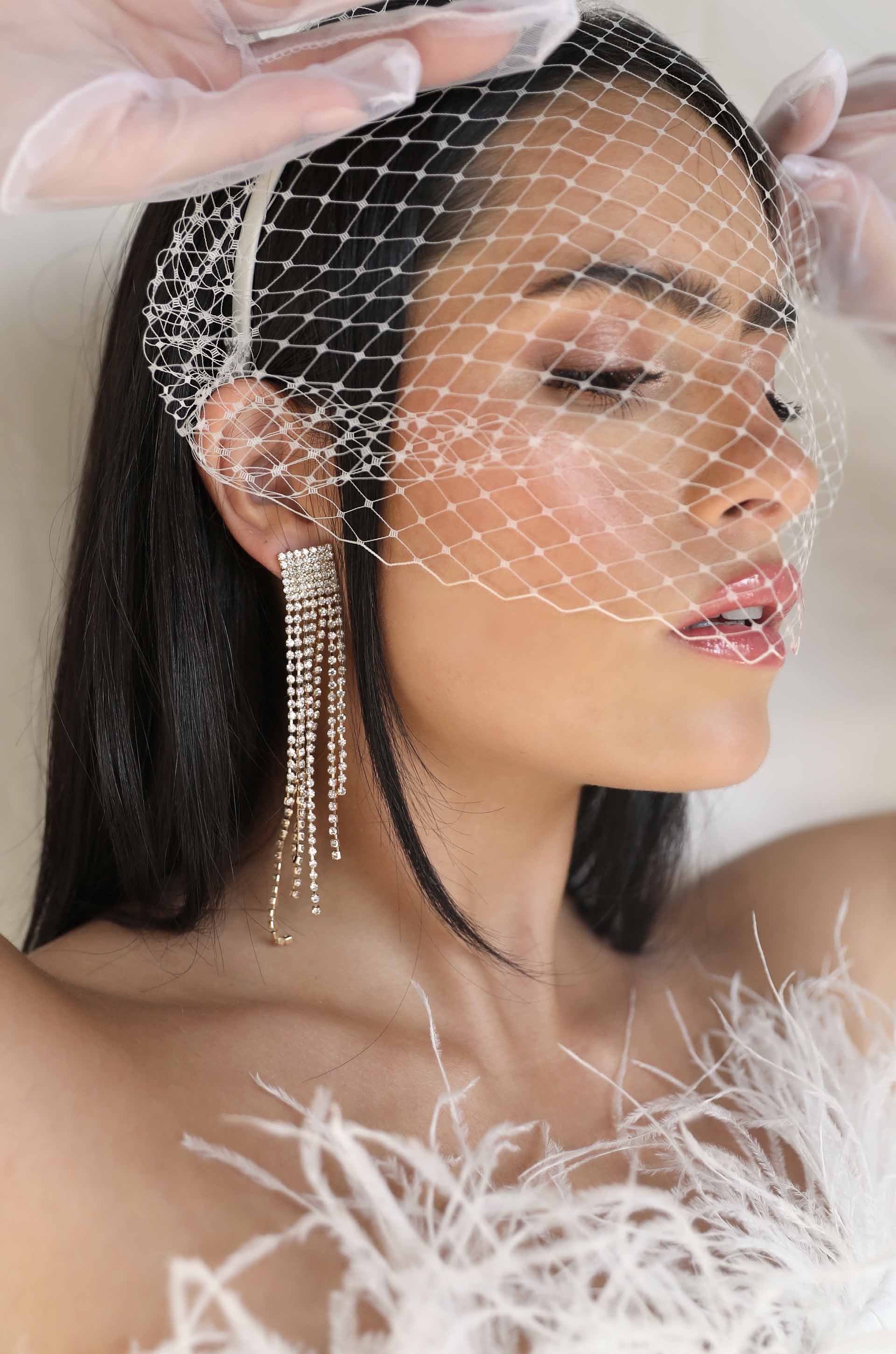 A woman with long, dark hair wears an intricate veil and dangling earrings. She gazes softly while her hand adjusts the veil. Her outfit features delicate, feathery details.