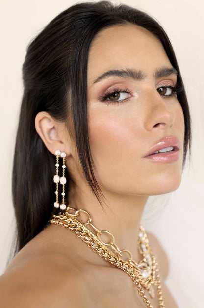 A woman with long, sleek black hair gazes confidently at the viewer. She wears elegant, multi-layered gold jewelry, complementing her glowy makeup, against a soft, neutral background.