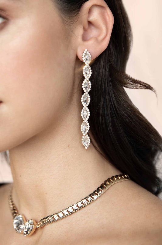 A long, sparkling crystal earring hangs from the ear of a woman, complementing a gold chain necklace with heart-shaped accents. The background is softly blurred.