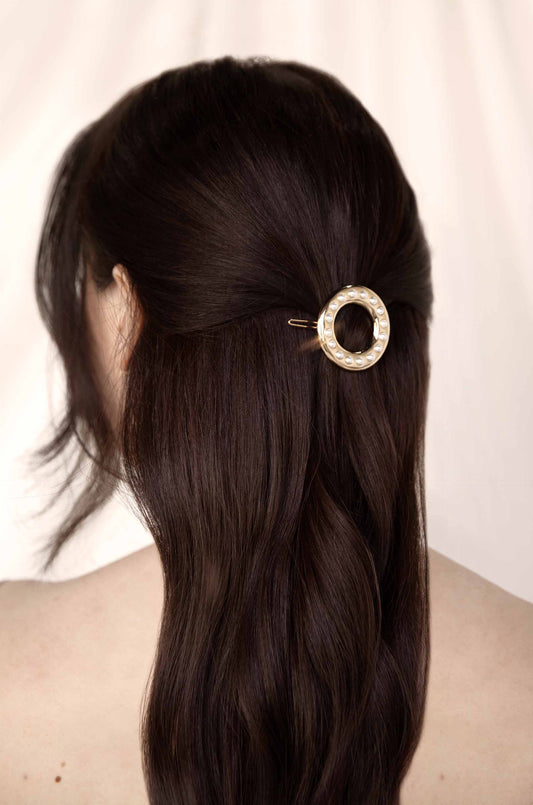 A hair clip, adorned with pearls, secures long, dark hair back from the face, revealing soft waves. The background is softly blurred, enhancing the focus on the hair and clip.