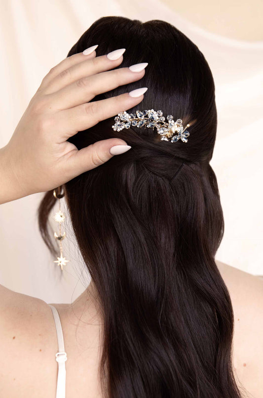 A hand gently adjusts a decorative hairpiece in dark, wavy hair. The background is a soft, neutral tone, enhancing the elegance of the hairstyle and accessory.