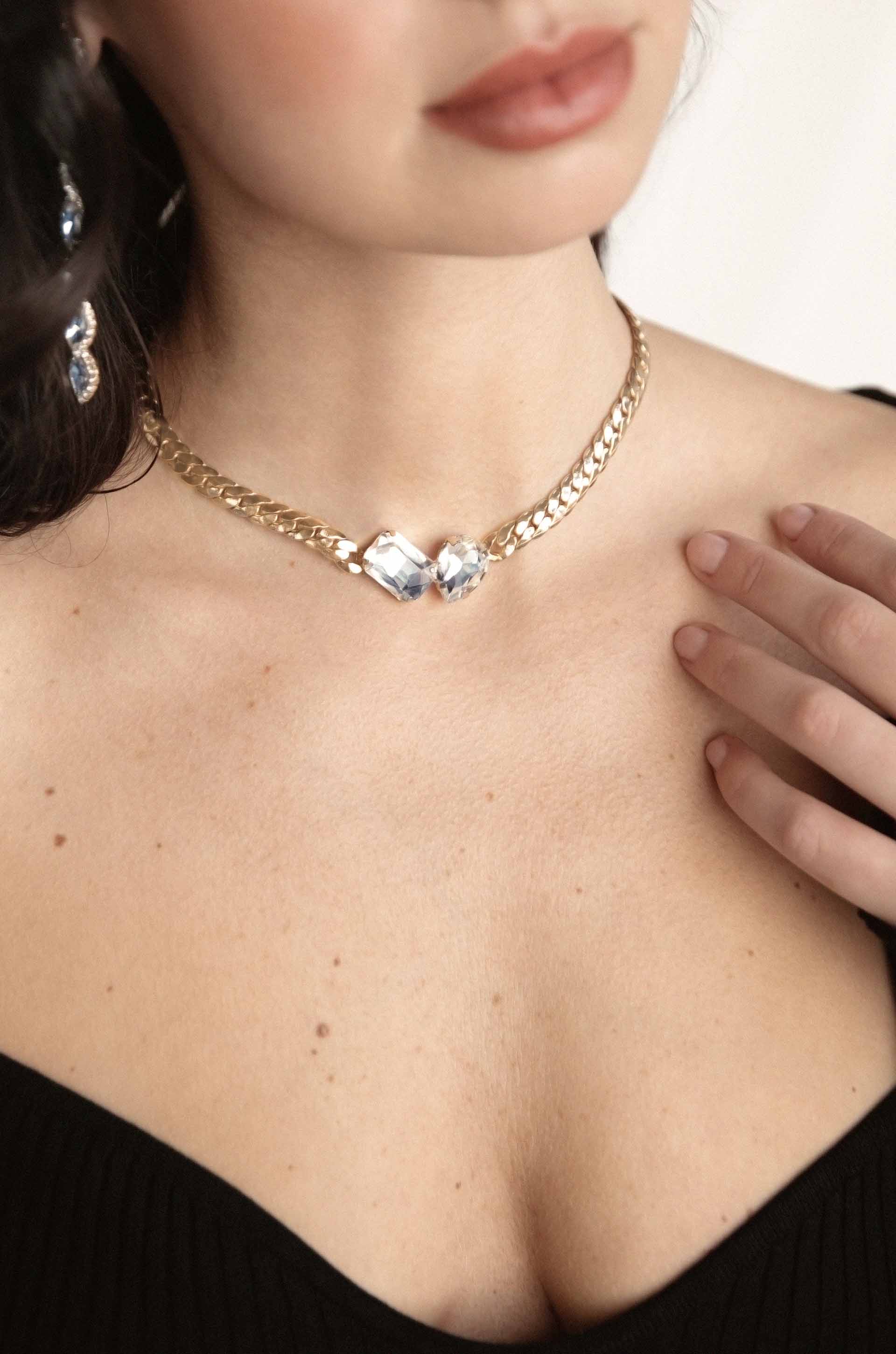 A close-up of a woman's neck adorned with a gold chain necklace featuring two large, clear crystals. She gently touches her collarbone, wearing a black top against a soft background.