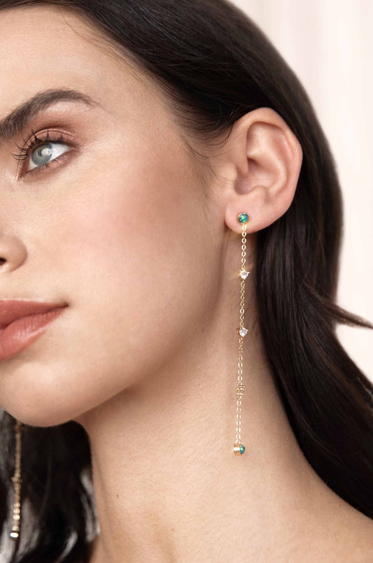 A close-up of a person's ear features a delicate gold earring with hanging chains and colorful embellishments. The soft background highlights the intricate jewelry design and the person's facial features.