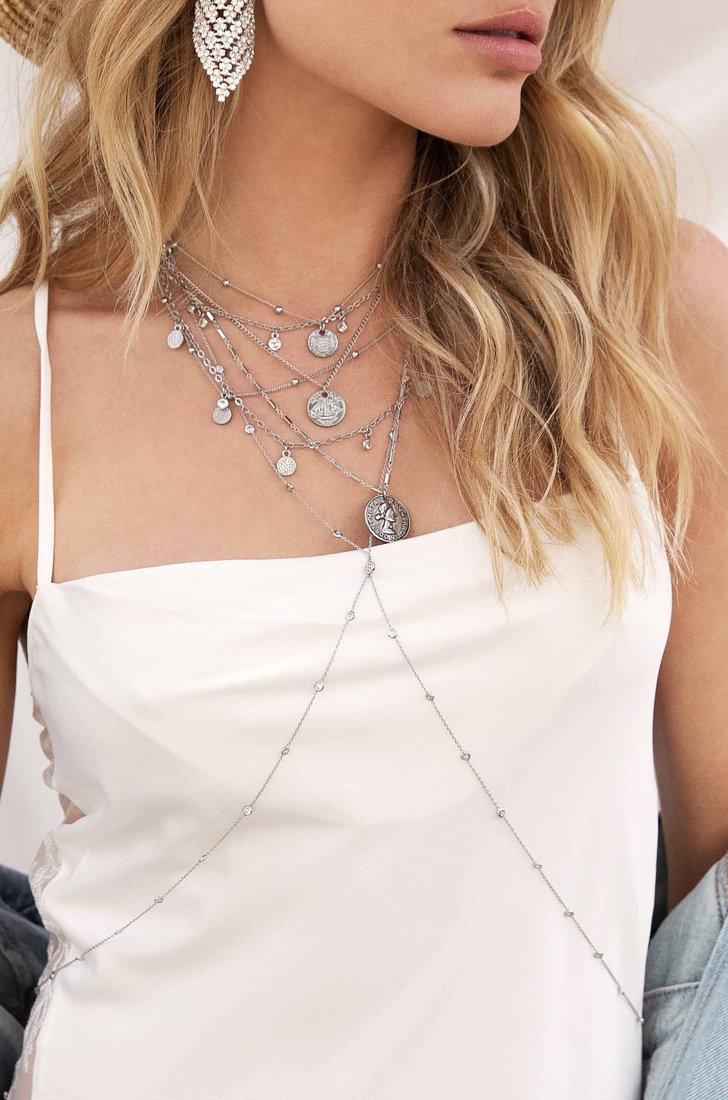 A woman wears multiple layered silver necklaces over a white camisole, showcasing intricate pendants. Her wavy hair frames her face, complemented by a large, sparkling earring.