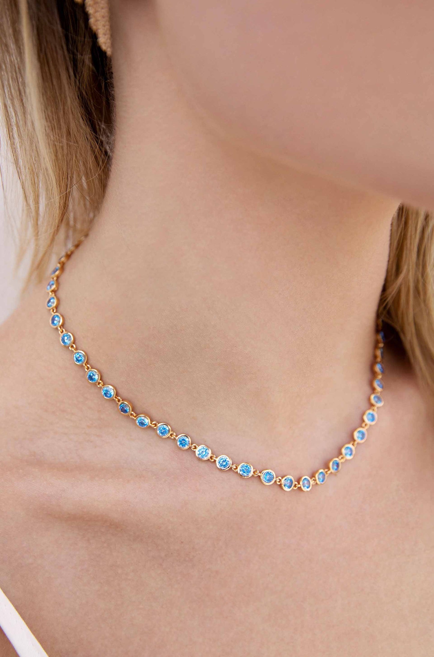 A gold necklace adorned with small blue gemstones rests on a person's neck, highlighting their skin tone and collarbone in a well-lit, neutral setting.