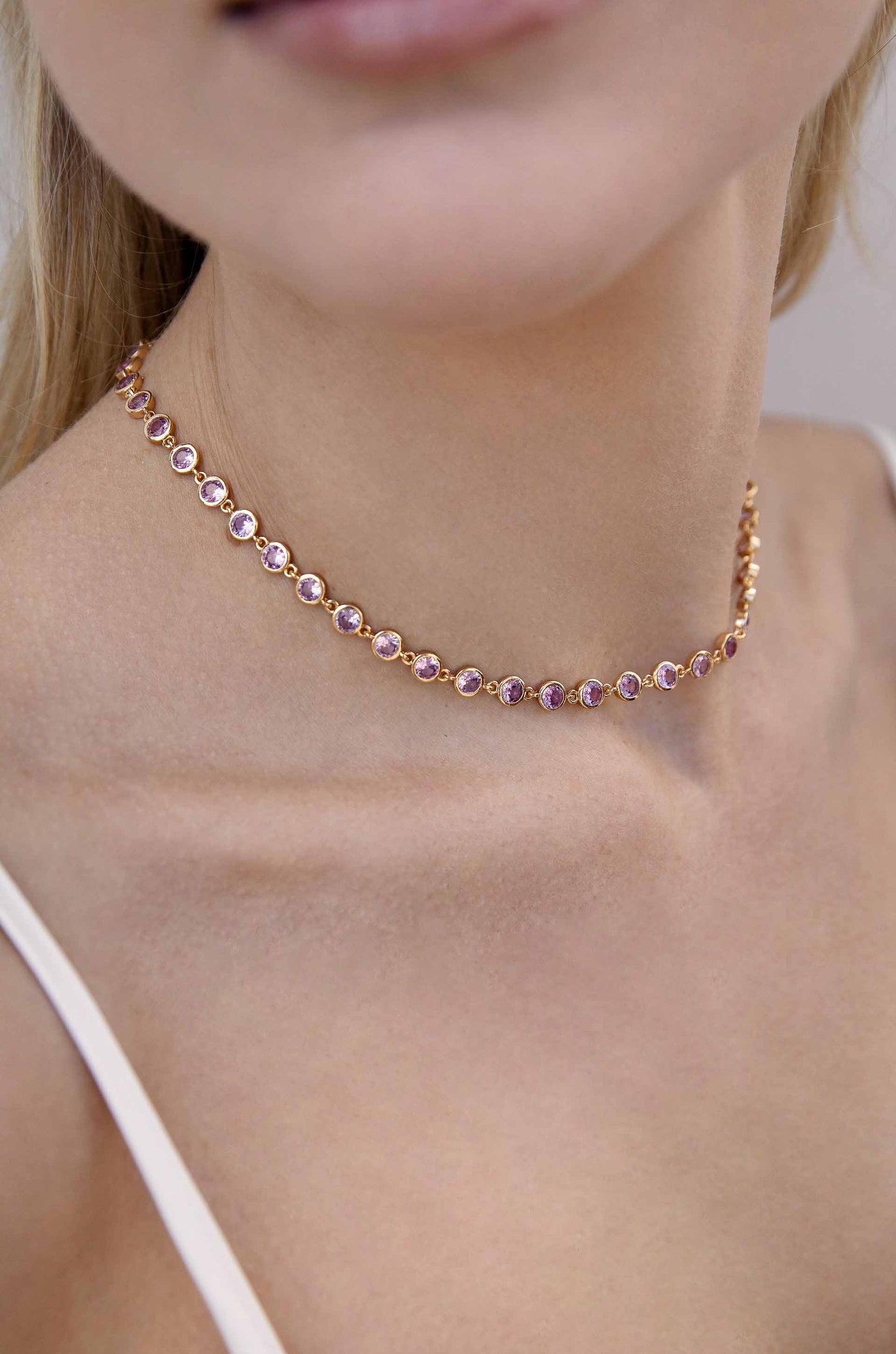 A delicate rose gold choker necklace adorned with round purple gemstones rests gracefully on a woman's neck, complemented by her smooth, bare skin in a softly lit environment.