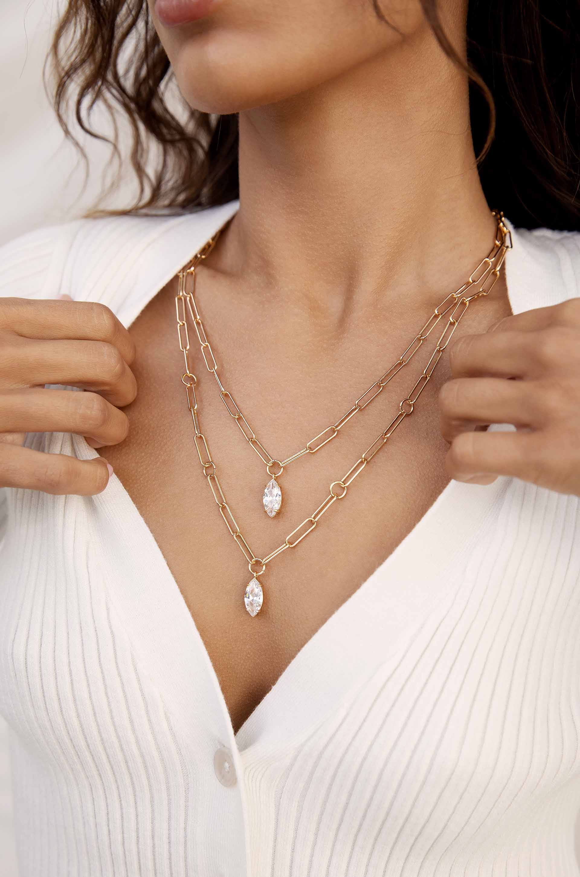 A woman is adjusting two layered gold necklaces with crystal pendants, wearing a white ribbed cardigan. The setting is softly lit, highlighting the jewelry against her skin.