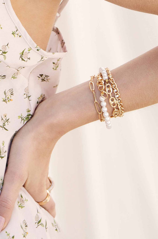 A wrist adorned with multiple bracelets, including gold chains and pearl accents, rests against a floral-patterned fabric, suggesting a casual yet stylish summer setting. The background appears soft and light.