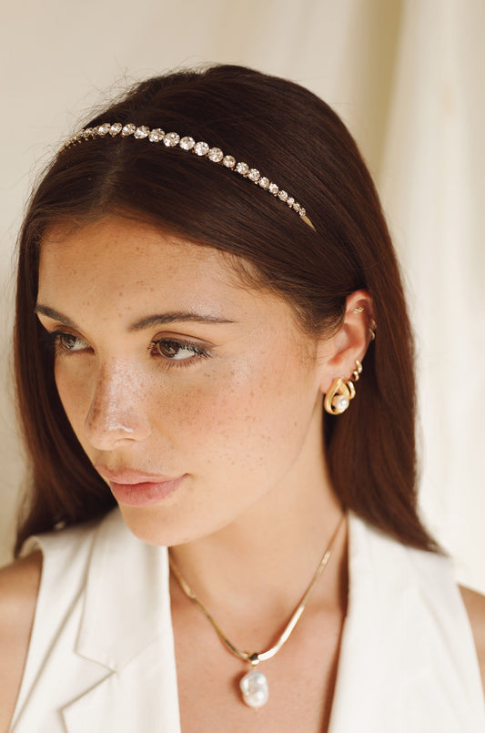 A young woman with long, straight brown hair wears a jeweled headband and elegant jewelry. She looks thoughtfully to one side against a soft, light-colored background.