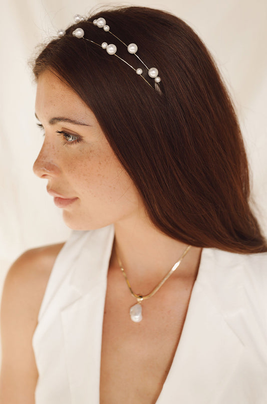 A young woman with long brown hair is wearing a pearl-encrusted headband and a gold necklace with a pendant. She poses against a soft, neutral background, showcasing her profile.