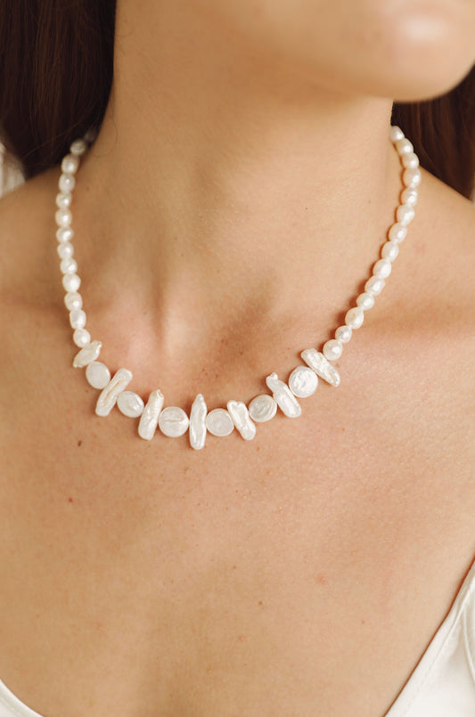 A pearl necklace adorns the neck of a person, showcasing a mix of round and irregularly shaped pearls against a smooth skin background, emphasizing elegance in a light, neutral context.