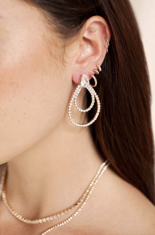 A close-up view of a woman's ear adorned with sparkling drop earrings and multiple ear cuffs. Soft, natural skin tones and long, dark hair provide a subtle, elegant backdrop.