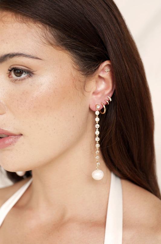 A woman with long, dark hair wears a delicate, dangling earring featuring pearls and small crystals. The soft background enhances the jewelry's elegance and the subtle freckles on her cheek.