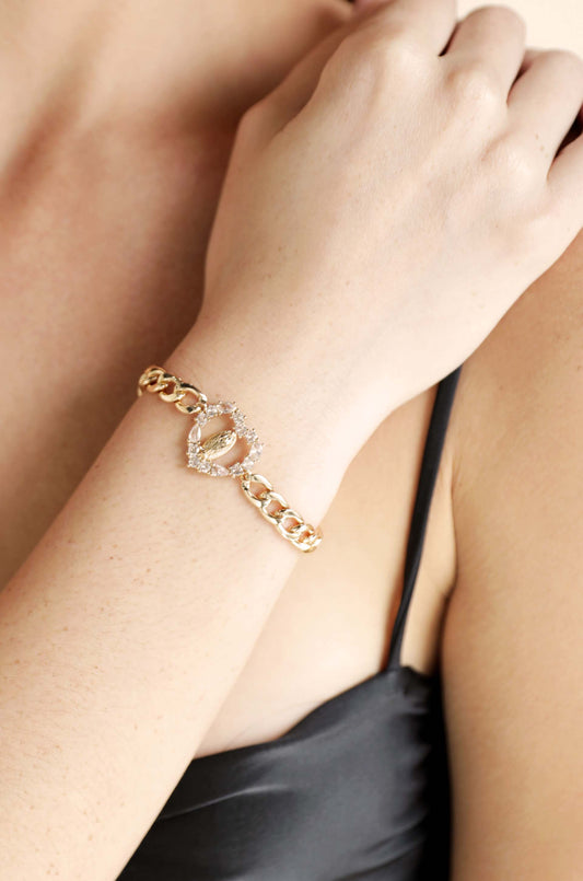 A gold bracelet with a heart-shaped clasp is draped around a woman's wrist, while she gently touches it with her other hand. The background features soft, neutral tones.