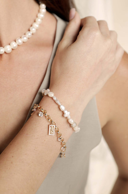 A close-up of a person's arm features two bracelets: one with white pearls and another in gold with decorative charms. The background is soft and neutral, enhancing the accessories' elegance.