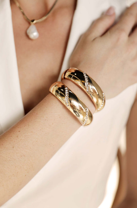 Two gold bracelets, adorned with diamonds, are worn on a wrist. The setting features a light-colored outfit and a pearl necklace, highlighting elegance and style in a close-up.