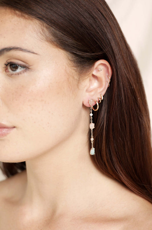 A close-up of a woman's ear adorned with a long, dangling earring featuring various stones and pearls, showcasing a subtle, natural beauty against a soft, neutral background.