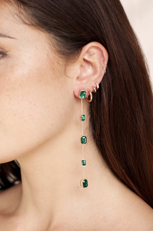 An elegant, long earring with four green gemstones hangs from a person's ear. The person has dark brown hair and a minimalist jewelry style, showcasing a polished look.