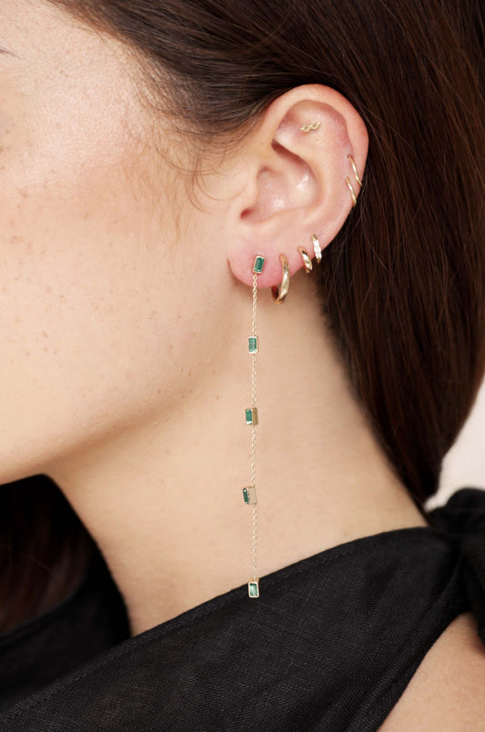 A woman's ear showcases multiple gold earrings, including a long, dangling earring with rectangular green stones, set against a backdrop of dark fabric. The skin tone reveals slight freckles.